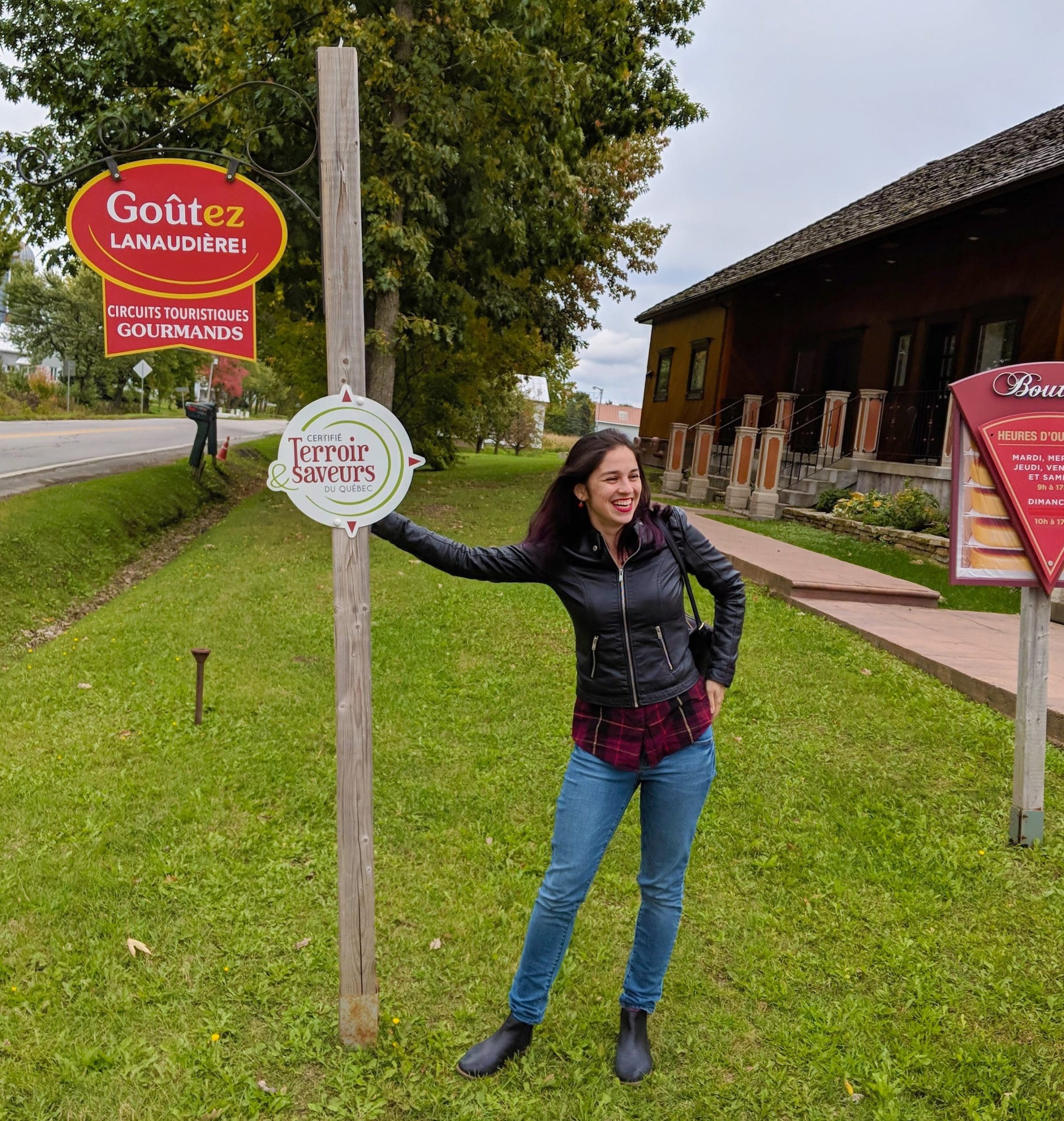 Eliane appuyée sur un poteau avec le panonceau de Terroir et Saveurs, devant la fromagerie Du Champ à la Meule