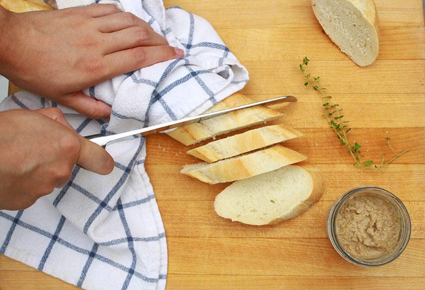 Une main tranche un pain sur une planche de bois, à côté d'un pot de mousse de foie gras d'oie ouvert